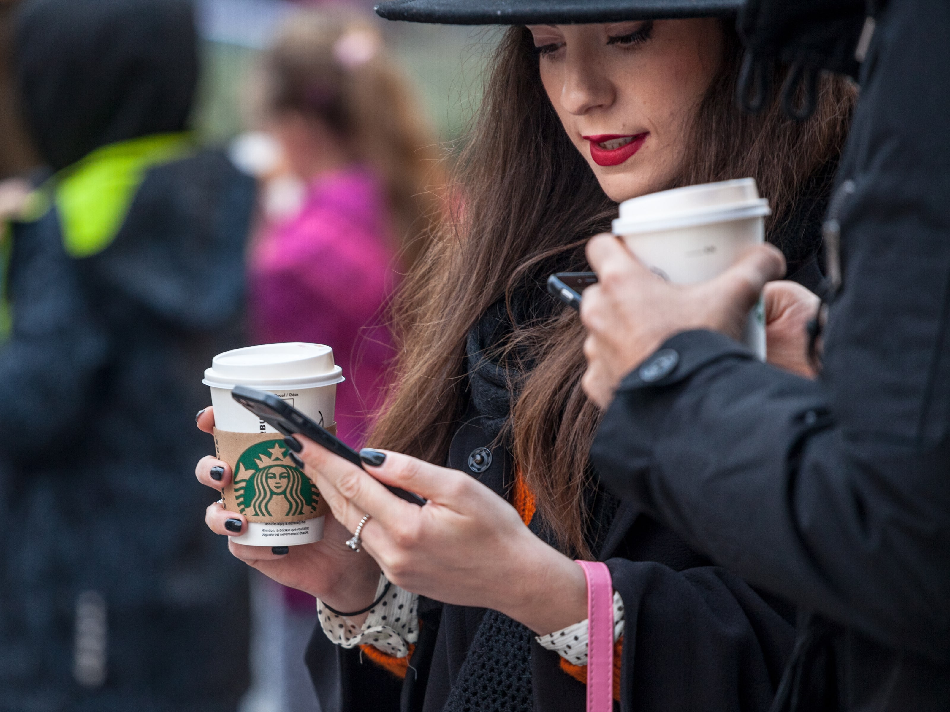Picture of a young pregnant woman holding her smartphone while drinking a coffee in a cardboard coffee cup with the logo of Starbucks. 